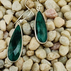 May include: A pair of teardrop-shaped earrings with faceted, deep green gemstones set in silver-colored metal. The earrings are displayed against a background of small, smooth, light-colored stones.