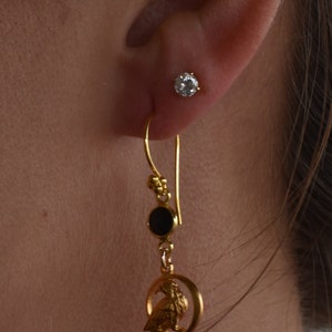 May include: Gold dangle earrings with a black stone, a small gold bird charm, and a gold hoop. The earrings are hanging from a person's ear.