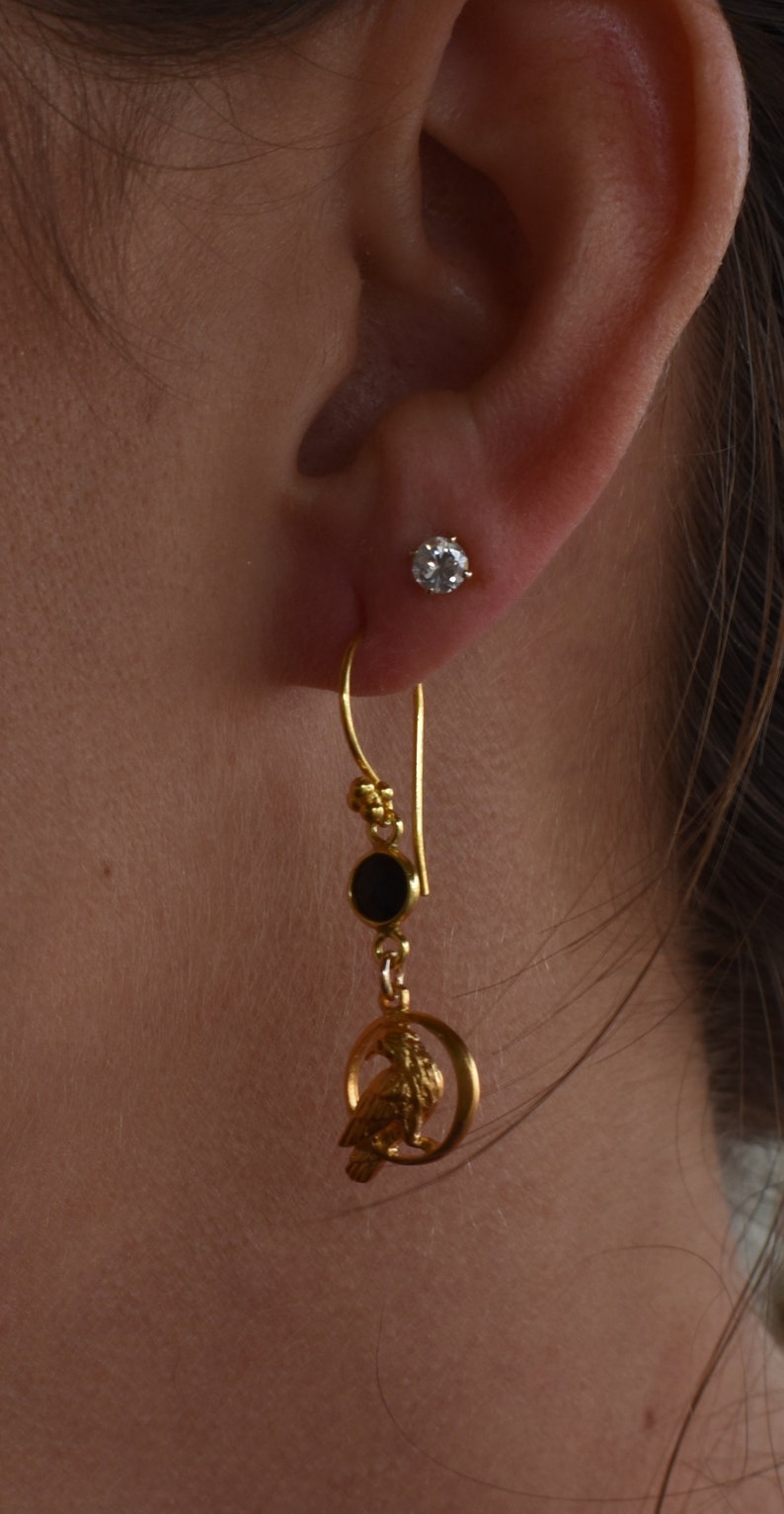 May include: Gold dangle earrings with a black stone, a small gold bird charm, and a gold hoop. The earrings are hanging from a person's ear.