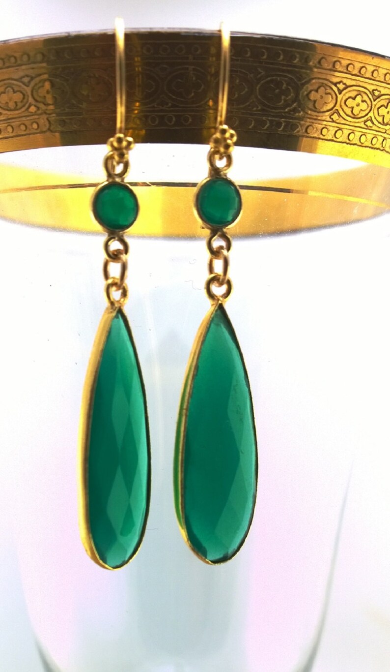May include: A pair of gold dangle earrings with green gemstone drops. The earrings have a teardrop shape and are accented with smaller green gemstones.