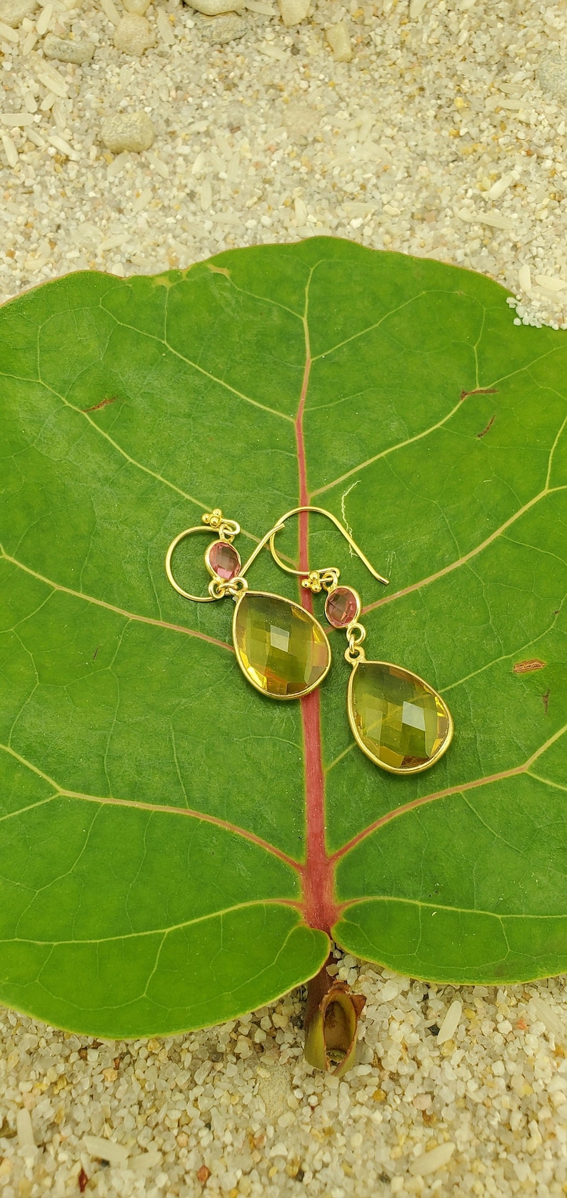 May include: Gold-tone drop earrings with yellow and pink gemstones. The earrings are displayed on a large green leaf with red veins, set against a sandy background. The earrings have a teardrop shape.