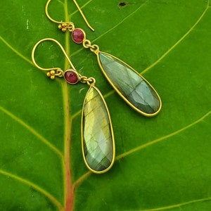 May include: A pair of gold dangle earrings with teardrop-shaped labradorite stones and small red gemstone accents.
