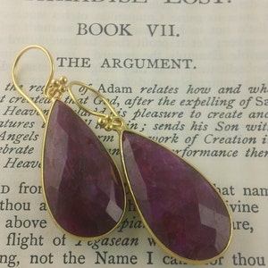 May include: A pair of teardrop-shaped earrings with gold settings and deep purple gemstone drops. The earrings are photographed on a book with the text "Paradise Lost" and "Book VII" visible.