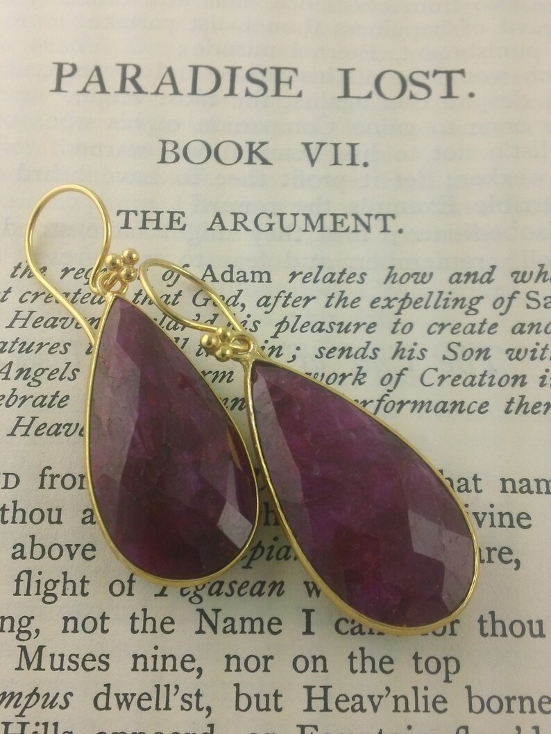 May include: A pair of teardrop-shaped earrings with gold settings and deep purple gemstone drops. The earrings are photographed on a book with the text "Paradise Lost" and "Book VII" visible.