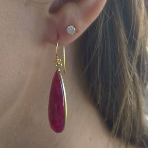 May include: A teardrop-shaped ruby gemstone earring set in a gold bezel with a gold hook wire, worn alongside a small round stud earring. The ruby is a deep, saturated red color with a faceted surface, suspended from a thin gold wire hook.