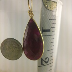 May include: A pair of gold earrings with a teardrop-shaped, faceted, deep red gemstone. The earrings are approximately 2 inches long.
