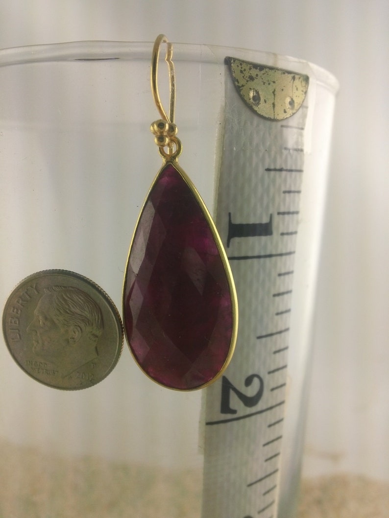 May include: A pair of gold earrings with a teardrop-shaped, faceted, deep red gemstone. The earrings are approximately 2 inches long.