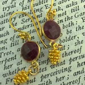 May include: A pair of gold earrings with a dangling charm. Each earring features a round, dark red gemstone and a gold charm that resembles a pine cone.