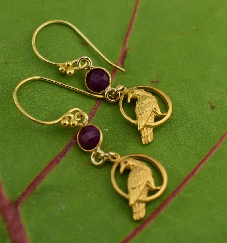 May include: Gold earrings with a purple gemstone and a detailed raven design.
