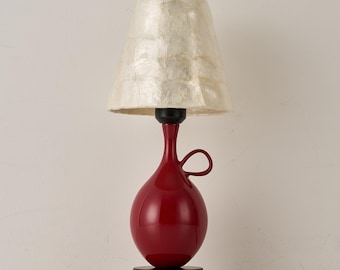 May include: A table lamp featuring a glossy red, bulbous ceramic base with a small side handle, a black circular base, and a textured, off-white, translucent conical lampshade. The lamp includes a black power cord extending from the base.