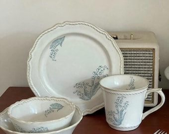 May include: A ceramic dinnerware set featuring a cream-coloured plate, two nested bowls, and a mug, all decorated with a blue-green lily of the valley floral pattern and a rope-textured rim detail. A metal fork with a wooden handle lies nearby.