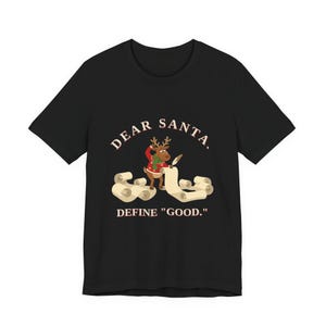 Op de afbeelding: Een zwart T-shirt met korte mouwen en een afbeelding van een bruin rendier met een rode trui en groene sjaal, schrijvend op een lange rol papier met een ganzenveer. De tekst luidt: DEAR SANTA, DEFINE "GOOD." Het ontwerp staat op de borst.