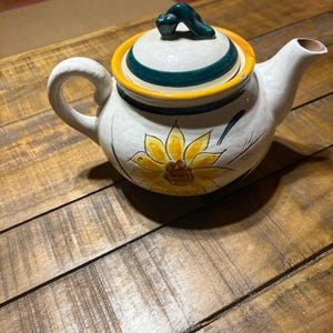 May include: A ceramic teapot with a white base, featuring a hand-painted yellow sunflower design on the front. The lid has a dark green handle and a yellow rim. The teapot includes a curved handle and a spout, with dark green accents throughout.