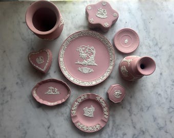 May include: A collection of pink jasperware ceramic pieces featuring white neoclassical relief decorations. The set includes a vase, a heart-shaped box, a flower-shaped box, a round plate, a small dish, a pitcher, an oval tray, and a round ashtray.