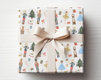 May include: A white gift box with a beige ribbon and bow. The box is wrapped in paper with a repeating pattern of nutcrackers, ballerinas, Christmas trees, and other holiday imagery.