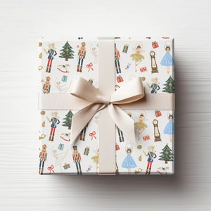 May include: A white gift box with a beige ribbon and bow. The box is wrapped in paper with a repeating pattern of nutcrackers, ballerinas, Christmas trees, and other holiday imagery.