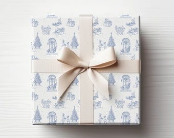 May include: A white gift box with a blue and white patterned design featuring illustrations of Christmas trees, houses, and people. The box is tied with a white satin ribbon and bow.