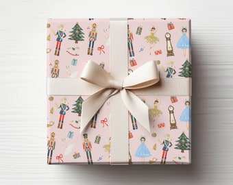 May include: A wrapped gift box with a light pink background featuring a pattern of nutcrackers, Christmas trees, ballerinas, and clocks. The box is tied with a cream-colored ribbon and bow, creating a festive and elegant presentation.
