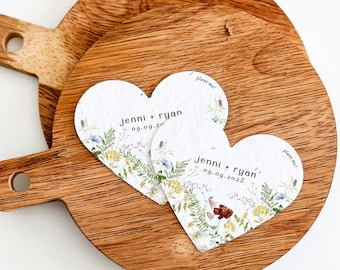 May include: Two heart-shaped paper cards with a wildflower print, the text "jenni + ryan" and "09.09.2025", and the phrase "plant me!" printed on them. The cards are placed on top of two round wooden boards with handles, featuring a visible wood grain.