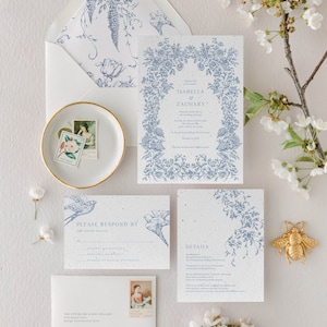 May include: A wedding stationery set featuring white paper with blue floral illustrations and text. Includes an invitation, RSVP card, details card, and envelopes. A small white dish holds two postage stamps, and a gold bee figurine sits nearby.