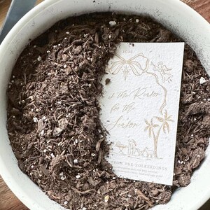 May include: A white round planter filled with dark brown potting soil and a rectangular off-white card featuring gold line art of a bow, an angel, palm trees, and a nativity scene with the text "He is the Reason for the Season" and 2023.
