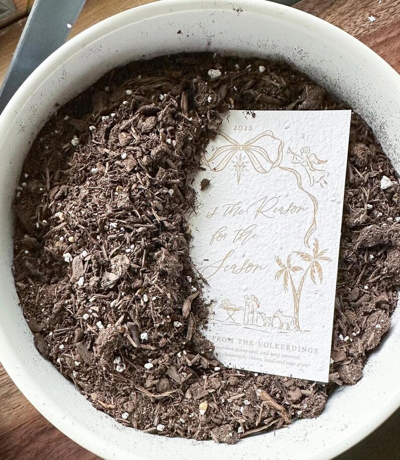May include: A white round planter filled with dark brown potting soil and a rectangular off-white card featuring gold line art of a bow, an angel, palm trees, and a nativity scene with the text "He is the Reason for the Season" and 2023.