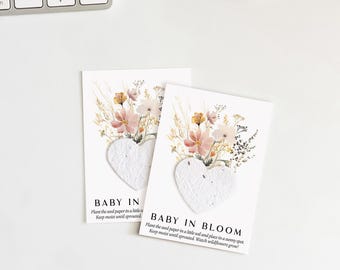 May include: Two white cards with a heart-shaped seed paper and floral arrangement. The text "BABY IN BLOOM" is printed on the cards, along with instructions to plant the seed paper. A keyboard and mouse are in the background.