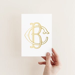 May include: A white card featuring a gold monogram with the letters "B" and "C" intertwined in an elegant, vintage style. The monogram is centered on the card, with a decorative flourish. The card is held by a hand.