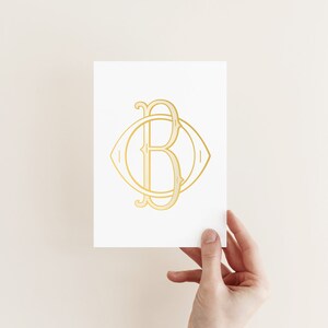 Puede incluir: Una tarjeta blanca con un monograma dorado de las letras "C" y "B" entrelazadas dentro de un círculo. El monograma es de estilo vintage.