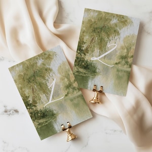 May include: Two rectangular table number cards featuring an impressionistic painting of a green willow tree by a river. The card on the left displays the white number 1, and the card on the right displays the white number 2. Each card is held by a gold stand.