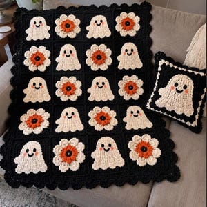 May include: A crochet blanket and matching square cushion featuring a repeating pattern of cream-coloured ghosts with pink cheeks and orange-centred flowers, all set against a black background with a scalloped black border.
