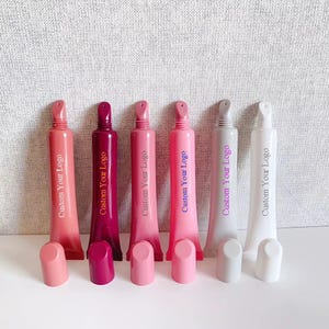May include: Six squeeze tubes for lip gloss or cosmetic products, each featuring an angled applicator tip and a removable cap. The tubes are colored in shades of coral, dark pink, light pink, bright pink, light gray, and white, with the text Custom Your Logo printed vertically on each.