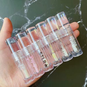 May include: Six clear plastic lip gloss tubes with white printed text and caps. The tubes are labeled with various brand names including Stay Glossy, Popgloss, STLR, Danae by Mae, Simones Cosmetics, and Gloss Tinted, with 2.5ml or 2ml capacities.