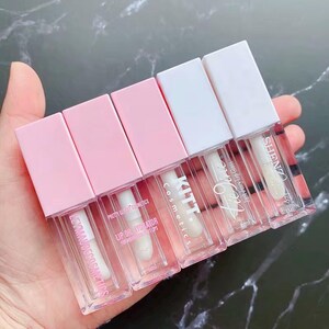 May include: Five rectangular lip gloss tubes with clear bodies and square caps. Three tubes have light pink caps and two have white caps. Each tube is labeled with brand names including KITT Cosmetics and SHENZ, and indicates a 6ml volume.