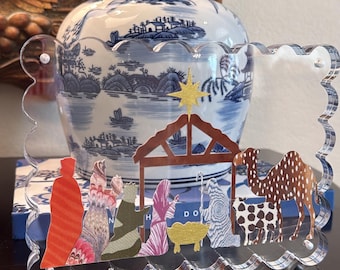 May include: A clear acrylic plaque with a scalloped edge featuring a colourful nativity scene. The design includes a gold star, a brown stable, a yellow manger, and various figures in patterned clothing, including a camel and a sheep.