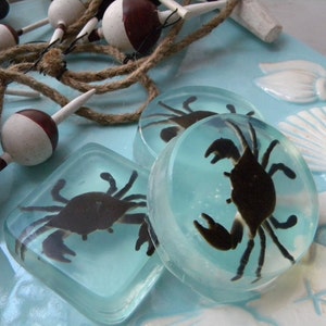 May include: Three clear resin coasters with a blue background and a black crab silhouette inside each. The coasters are on a blue and white background with seashells and rope.