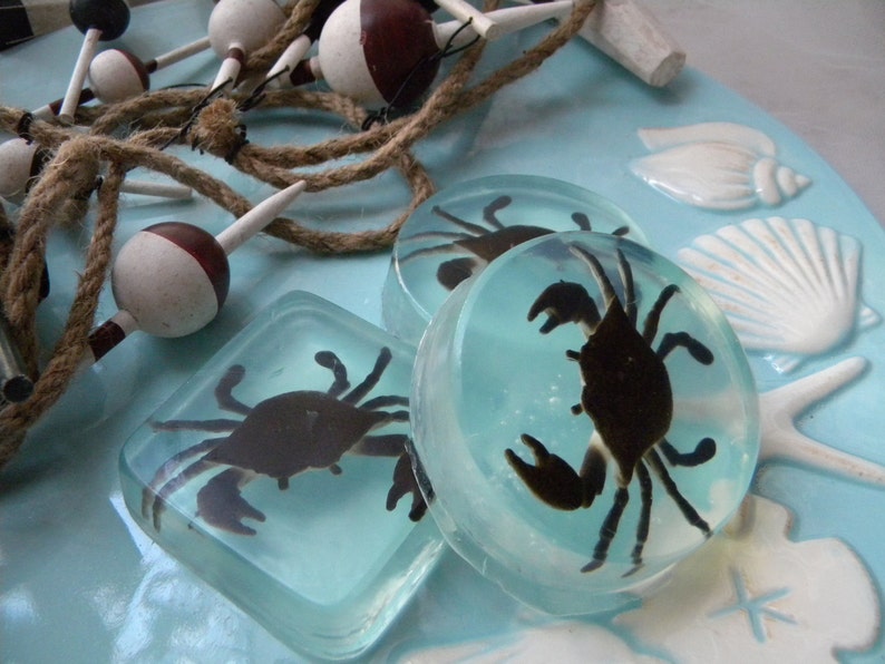 May include: Three clear resin coasters with a blue background and a black crab silhouette inside each. The coasters are on a blue and white background with seashells and rope.