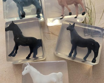 May include: Five horse figurines in clear, square blocks. The horses are in various colors: blue, pink, black, and white. The figurines are standing in a clear, gel-like substance.