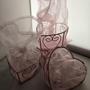 May include: Three decorative metal baskets in a soft pink hue, each with a heart shape or rounded design. The baskets are adorned with delicate, sheer pink fabric, creating a romantic and airy aesthetic. The metalwork features decorative swirls.