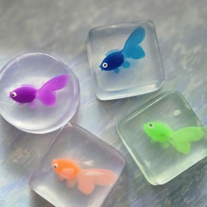 May include: Four handmade soaps with colorful fish designs. The soaps are clear and shaped as a circle and three squares. The fish are purple, blue, orange, and green. The soaps are a unique decorative item.
