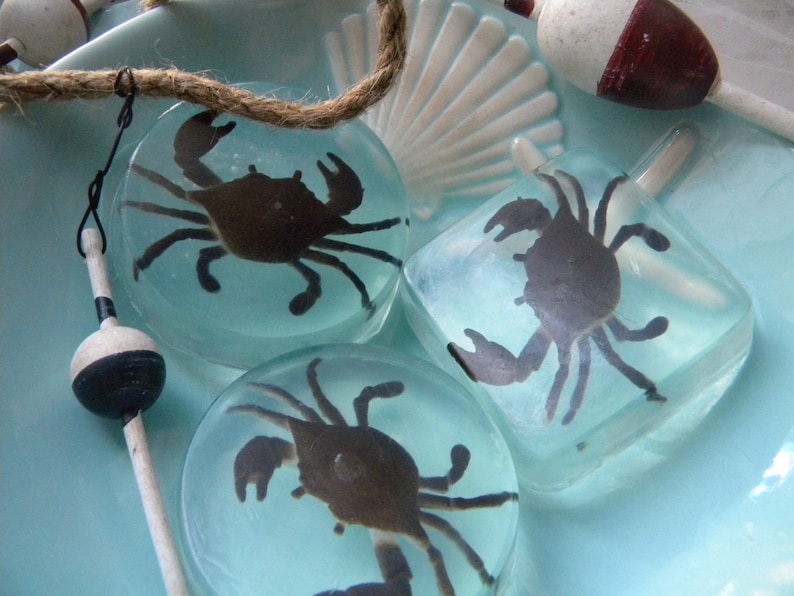 May include: Three clear resin coasters with a silhouette of a crab inside each. The coasters are on a blue and white background.