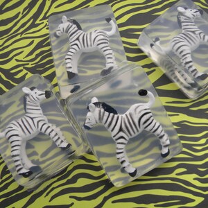 May include: Four clear soap bars with zebra figurines inside. The soap bars are on a black and yellow zebra print background.