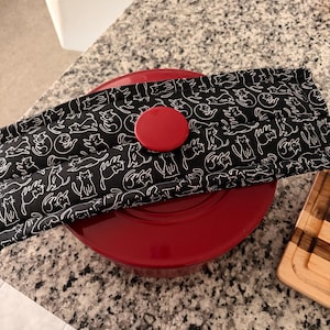 May include: A red round pot lid topped with a bright red knob, covered by a rectangular fabric handle cover featuring a black background with a repeating white line-art pattern of various cats in different sitting and playing poses.