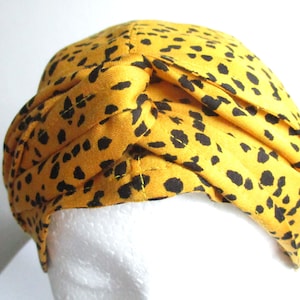Pode incluir: Um turbante amarelo com um padr&atilde;o de leopardo preto. O turbante &eacute; feito de tecido e &eacute; dobrado para criar um n&oacute; no topo.