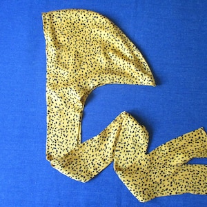 Pode incluir: Um turbante de cetim amarelo e preto com padr&atilde;o. O turbante &eacute; dobrado e amarrado no topo da cabe&ccedil;a.