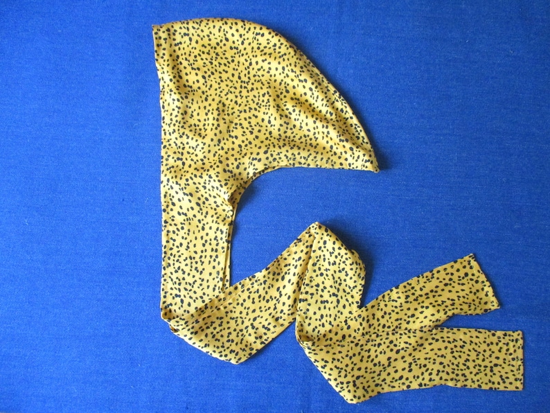 Pode incluir: Um turbante de cetim amarelo e preto com padr&atilde;o. O turbante &eacute; dobrado e amarrado no topo da cabe&ccedil;a.