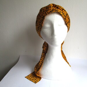 Pode incluir: Um turbante amarelo com bolinhas pretas. O turbante &eacute; amarrado com uma longa echarpe fluida que combina com o turbante.