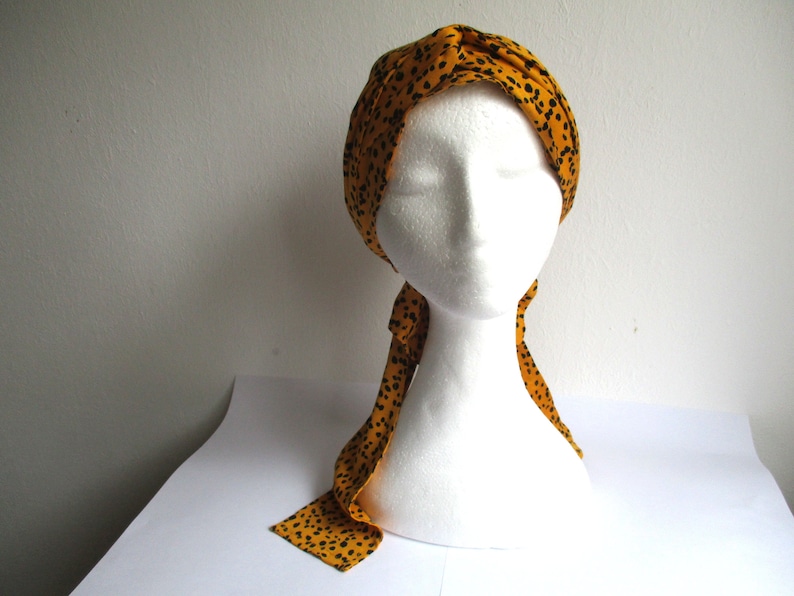 Pode incluir: Um turbante amarelo com bolinhas pretas. O turbante &eacute; amarrado com uma longa echarpe fluida que combina com o turbante.
