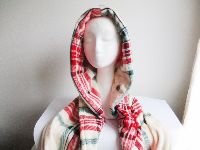 May include: A hooded scarf in red, white and green plaid. The scarf is made of a soft, fuzzy material and is worn around a white mannequin head.