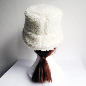 Puede incluir: Un gorro blanco y peludo de estilo bucket con una coleta marr&oacute;n que sobresale por debajo.